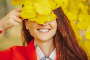 Woman with long brown hair in orange coat smiling and holding fall leaves in front of her face